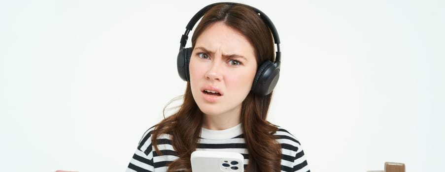 Portrait of girl in headphones with smartphone, looks confused at camera, frowns with perplexed face, stands over white background.