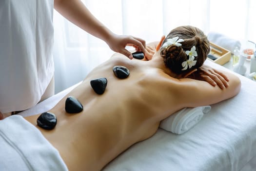 Hot stone massage at spa salon in luxury resort with day light serenity ambient, blissful woman customer enjoying spa basalt stone massage glide over body with soothing warmth. Quiescent