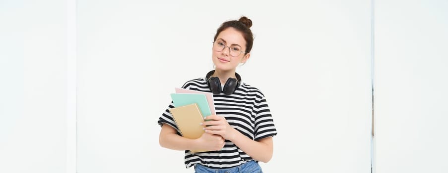 Student and education concept. Young woman with books, notes and pen standing over white background, college girl with headphones over neck posing in studio.