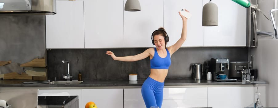 Portrait of beautiful sports woman, wearing activewear, dancing in kitchen with headphones, listening music and having fun, feeling energy after workout session.