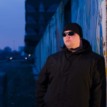 Upper body shot of middle-aged man in black jacket,cap and sunglasses leaning against a wall at dusk and looking to the side