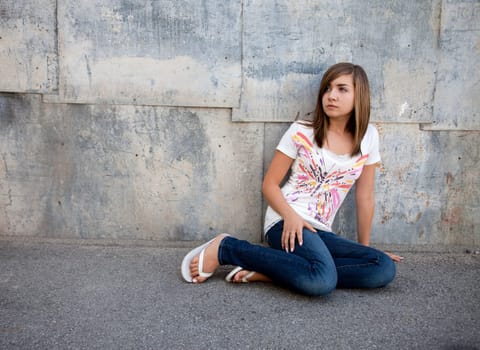 Outdoor photo of pretty teenage girl seated on pavement against grunge wall.