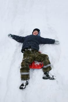 A cute 5-year-old boy in a blue winter jacket and a blue hat lies on the snow along with a small red sled.