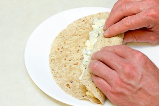 Two hands rolling up an egg salad and jalapeno mixture in a multi grain tortilla wrap on a white plate for breakfast.