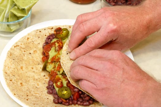 Two hands rolling up in a soft multi grain tortilla adzuki beans, salsa, guacamole, and tamed jalapenos for lunch.