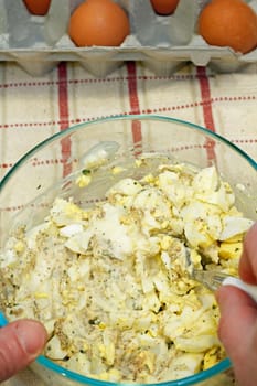 Two hands mixing egg salad mixture in a glass bowl with four raw brown organic eggs sitting in a cardboard carton on a towel on the counter.