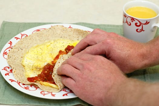 Two hands wrapping up two cooked eggs, tomato salsa, and black pepper in a multi grain soft tortilla with a cup of orange juice on the side.