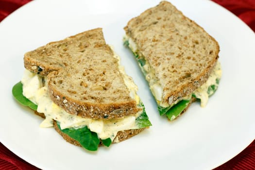 Egg salad sandwich with spinach leaves on a whole wheat grain with various seeds bread on a white plate on a red place mat.