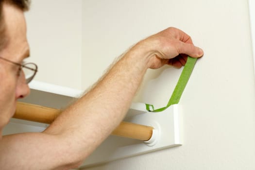 Having finished painting the walls of a closet a man removes the green painter&#039 s tape from around the shelf he was protecting.