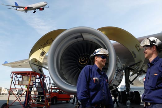 flight engineers, mechanics with large jet engine in background, airliner coming in for landing