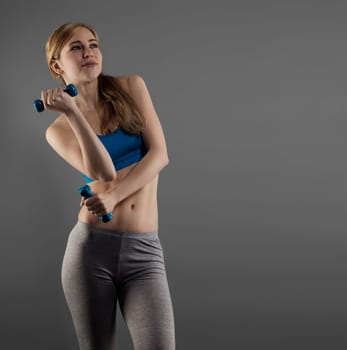 Young sportswoman with fitness dumbbells against gray background