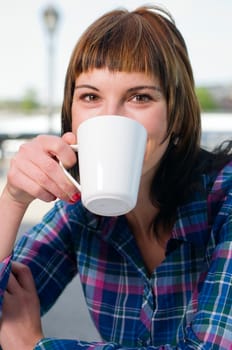 Head and shoulders shot of a young brown-haired woman sitting at a table in a street café and holding a white cup