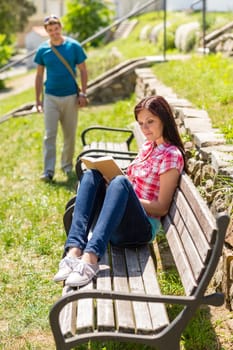 Young woman reading book on bench man coming towards her