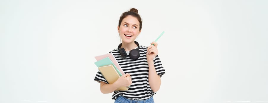 Portrait of young woman, student with notebooks and earphones on her neck, posing for college advertisement, white background.