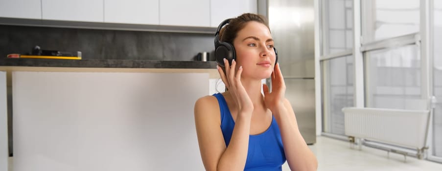 Young woman puts on wireless headphones to listen music during workout at home, wears sportsbra, smiles with pleased face.