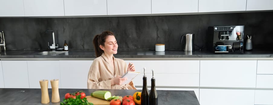 Portrait of woman thinking of menu, sitting in the kitchen and making grocery list, vegetables and meal ingredients on counter.