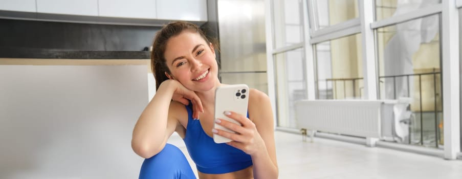 Young woman with smartphone does workout at home, using mobile phone app for sports training indoors, wears blue sportsbra and leggings.