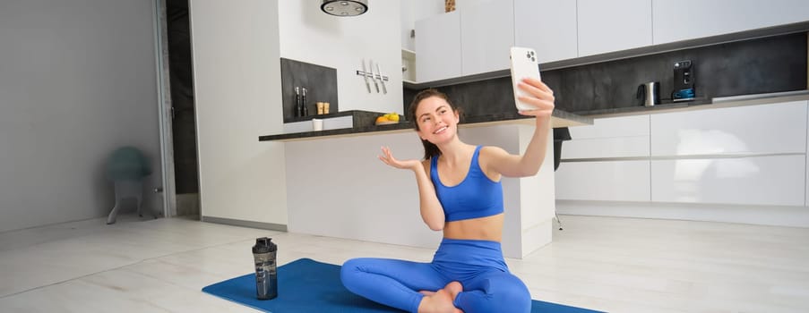 Active young woman does sports, records her workout training from home on smartphone camera, posing for selfie inside her house, sits on rubber yoga mat in blue leggings and sportsbra.