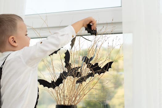 Halloween concept. Autumn holiday. The boy stands with his back near the window against the backdrop of a black vase with batting mice cut out of paper on dry branches. Close-up. Background.