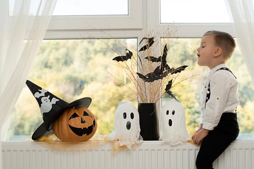 Halloween. Concept. A cute and cheerful boy sits near the window against the background of white ghosts and a vase with dry branches and black paper bats. Close-up.