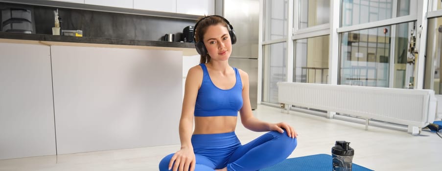 Young sporty athletic fitness trainer instructor woman wear blue tracksuit spend time in home gym use mobile phone headphones listen music. Workout sport concept.