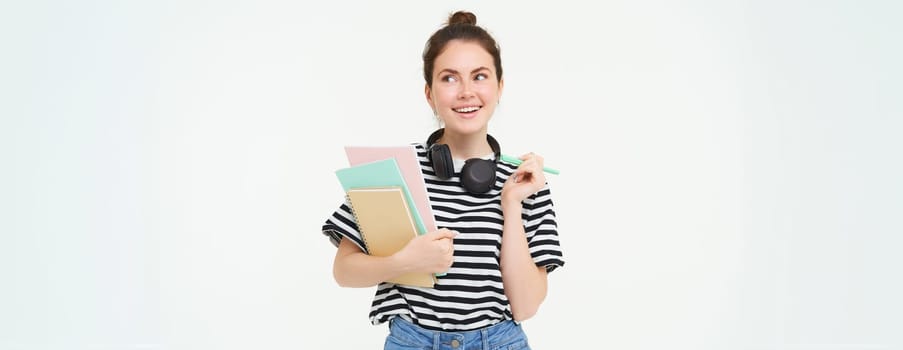 Portrait of young woman, student with notebooks and earphones on her neck, posing for college advertisement, white background.