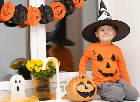 Halloween concept. A small, cheerful, handsome boy in a wizard's hat and an orange sweater sits on a table in the kitchen against the background of a pumpkin, a ghost and a bouquet of yellow flowers. Festive interior.