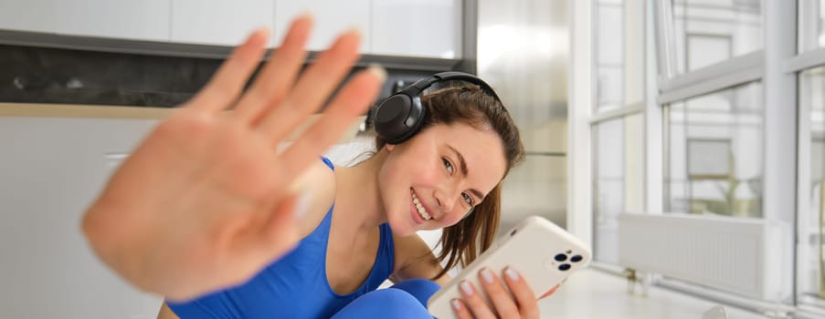 Portrait of beautiful brunette girl, hides from camera, puts her hand towards camera, listens music in headphones and holds smartphone, watches exercses online on mobile phone, smiles.
