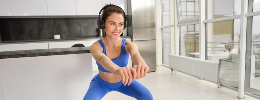 Image of young sportswoman, fitness instuctor doing squats workout at home, listening music in wireless headphones and stretching arms, doing exercises in living room.