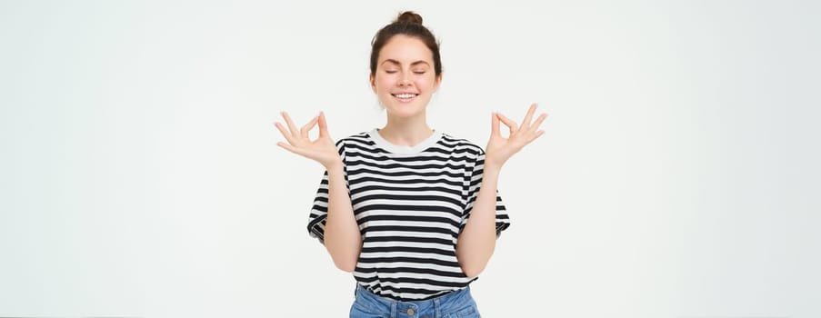 Portrait of young woman relaxing, feeling zen, meditating, smiling with pleased, calm face expression, standing over white background.