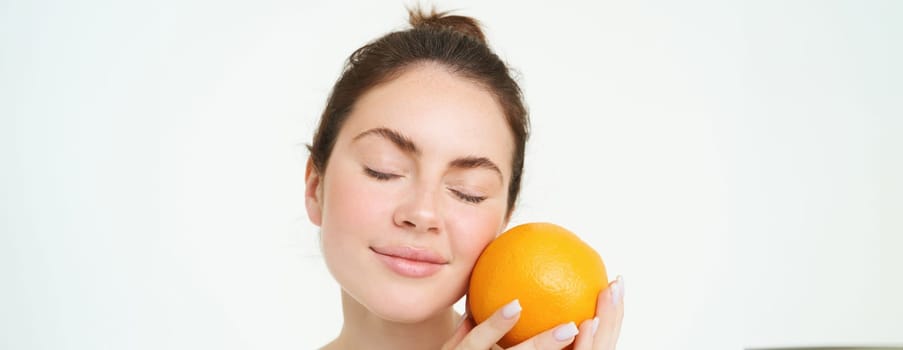 Portrait of young woman holding orange near face without blemishes, smiling, concept of vitamins, nourishment and organic facial treatments, white background.