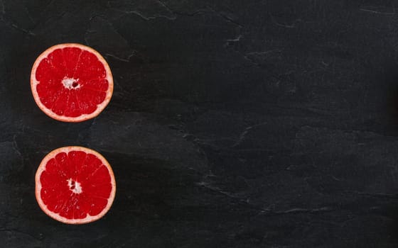 Pink grapefruit cut in half on black slate board photo from above. Wide banner with space for text right.