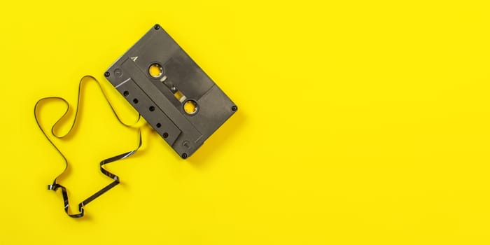 Tabletop view, audio cassette with some tape out on yellow background. Wide banner space for text right