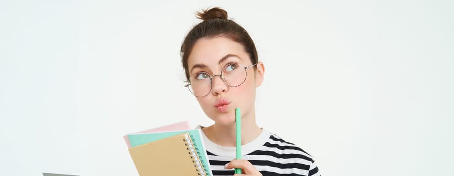 Portrait of cute woman, student in glasses, thinking, standing thoughtful with notebooks, memo notes, white background.