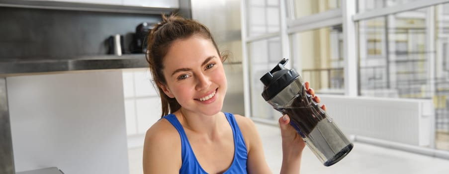 Smiling fitness girl drinks water in the middle of training session, smiling and looking happy, workout from home in blue sportsbra.