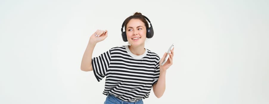 Young woman with smartphone listening to music, dancing to her favourite song in headphones, posing against white background.