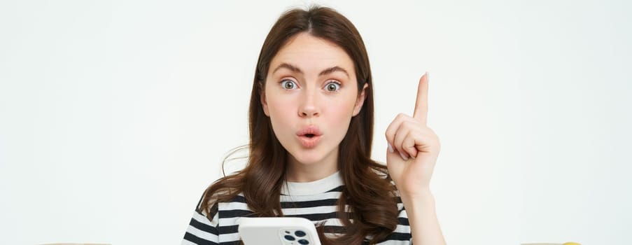 Portrait of excited woman raising finger, eureka gesture, has an idea or solution, holds mobile phone, white background.