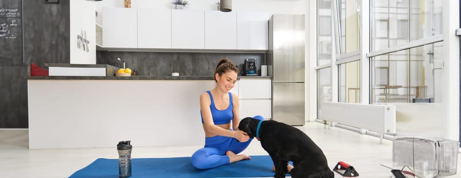 Portrait of beautiful young woman tries to do fitness workout from home, sits on floor on yoga mat with her cute dog, puppy wants to play while girl does exercises.