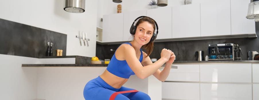 Portrait of active and healthy young woman, doing squats with resistance band, workout at home, listening music in wireless headphones.