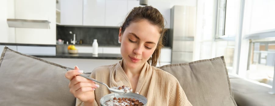 Image of smiling, happy young woman eating breakfast, holding bowl of cereals with milk, having meal at home.
