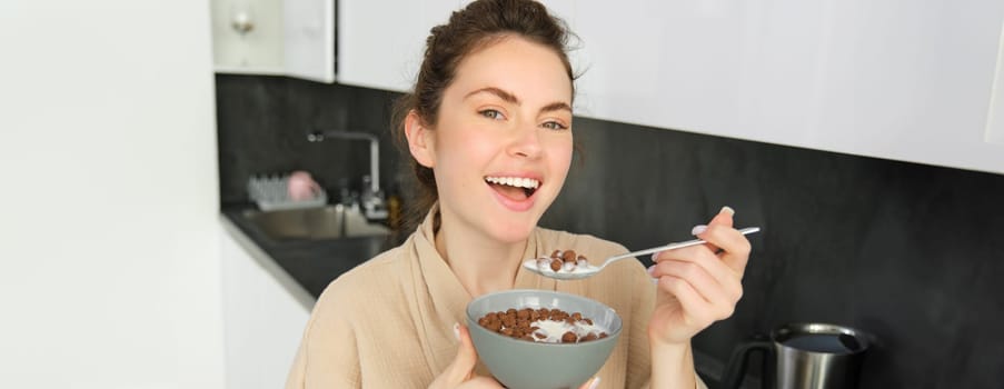 Happy mornings. Gorgeous young woman eating cereals with milk, standing in kitchen with breakfast bowl, enjoying start of the day, smiling.
