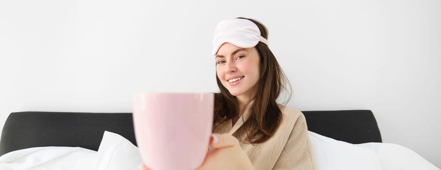 Beautiful smiling modern woman in pyjamas and sleeping mask, offering cup of tea or coffee in bed and looking happy, sitting in her bedroom and relaxing.