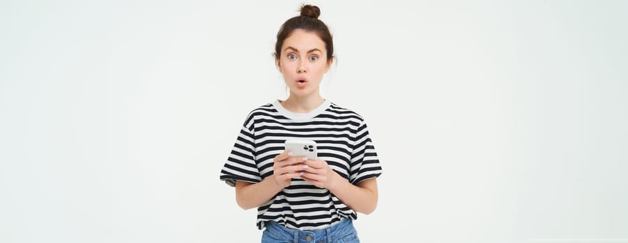 Portrait of impressed girl looks amazed, gasping, holding mobile phone, standing against white background. Copy space