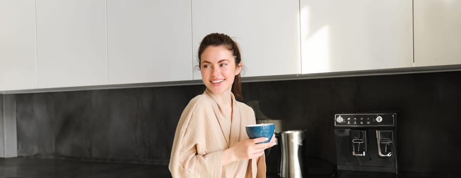 Portrait of beautiful housewife, young woman in bathrobe, holding cup of coffee, drinking tea in the kitchen, enjoying morning.