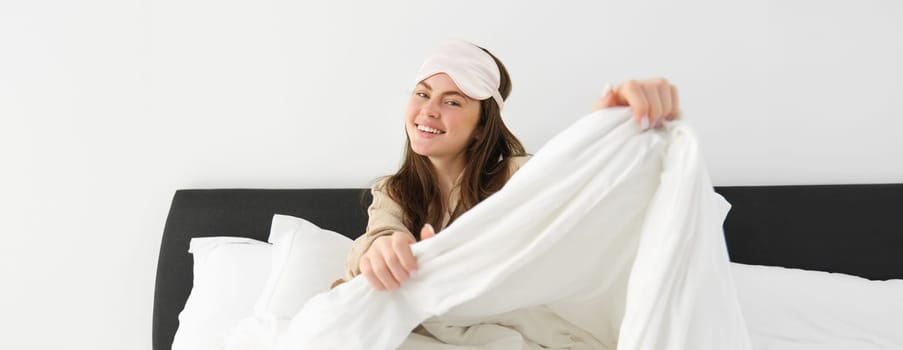 Beautiful young woman with sleeping mask on forehead, waking up in morning and smiling, resting in her bedroom, feeling lazy on weekends.