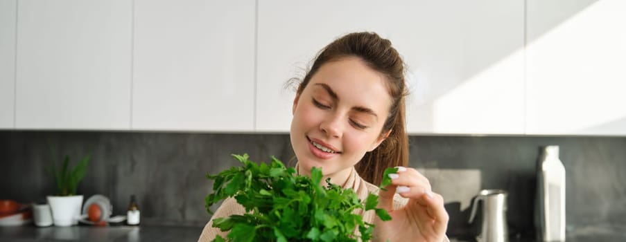 Portrait of beautiful smiling girl with bouquet of parsley, standing in the kitchen and cooking, adding herbs to healthy fresh salad or meal, preparing food.