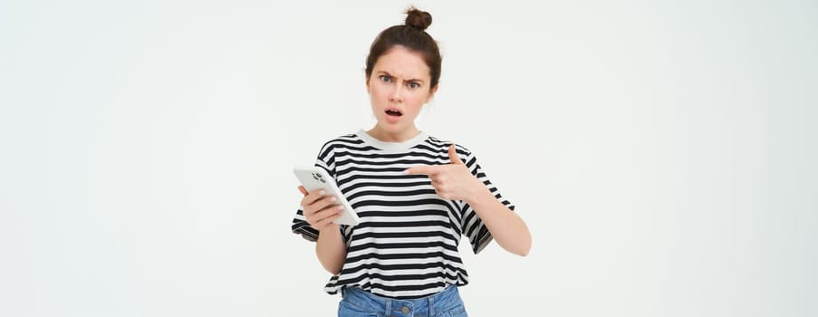 Image of angry young woman complains, points at telephone and looks disappointed, isolated against white background.
