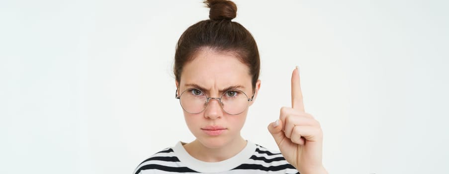 Close up portrait of angry, serious woman in glasses, pointing finger up, showing smth important, frowning with furious, confident expression, white background.