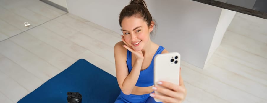 Portrait of young styling fitness girl doing workout from home, taking selfie and video for social media, gym instructor records her training session indoors.