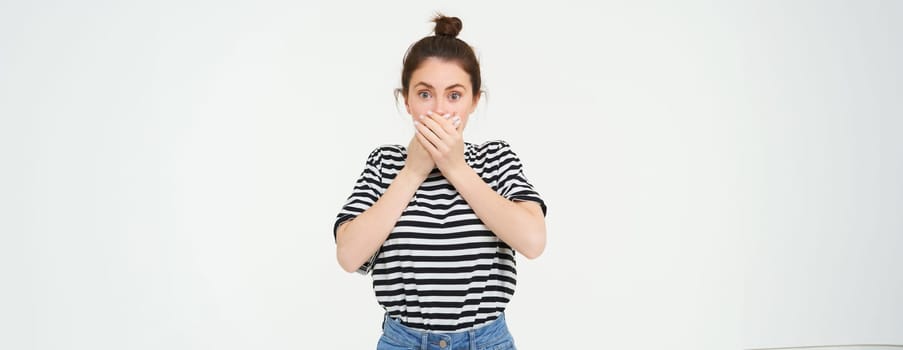 Portrait of woman looking shocked, covers her mouth, looks speechless and amazed, stares with disbalief, stands over white background.